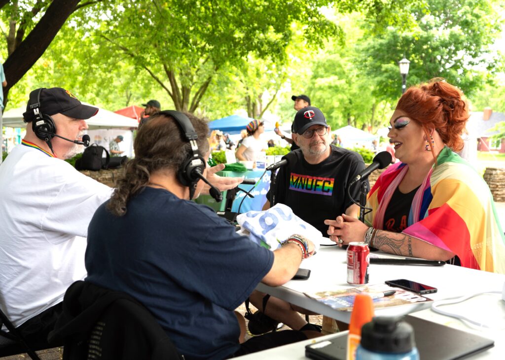 Radio hosts and a drag queen sit around a table, talking into microphones.