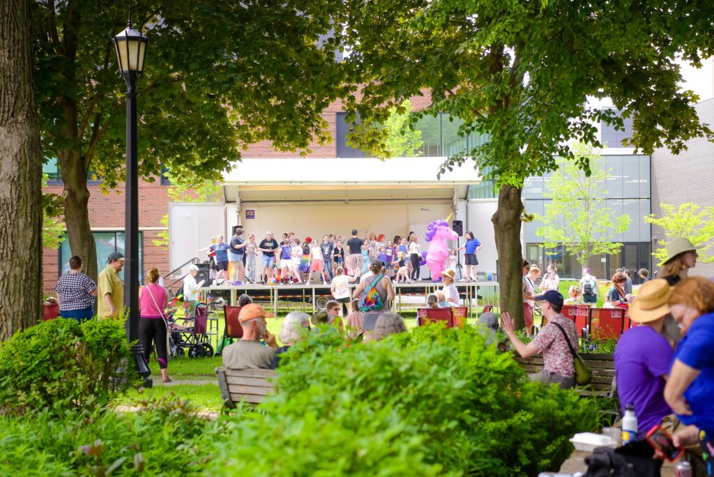 People gather on a stage and under trees in a park.